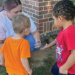 two-kids-looking-closely-at-a-frog-held-by-a-teacher-outside-a-brick-building