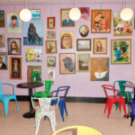 Framed-artwork-on-a-lilac-colored-wall-behind-several-tables-with-bold-color-metal-chairs-surrounding-them