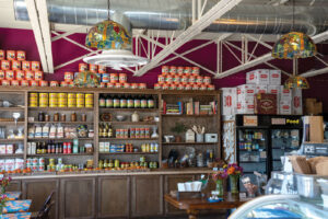 Step into a cozy store interior where wooden shelves brim with an array of canned goods and products. Colorful lamps hang above, casting a warm glow over the Stockyards experience. A refrigerated section beckons from one side, and a wooden table adorned with flowers completes the inviting scene.