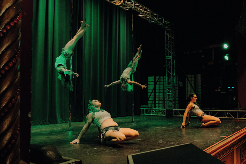 A group of four performers on stage captivate the audience with their pole dancing routine. Two are mid-spin, showcasing athletic poses that highlight the workout nature of their craft, while the others pose on the floor. The stage glows with green and yellow lights against a dark curtain backdrop.