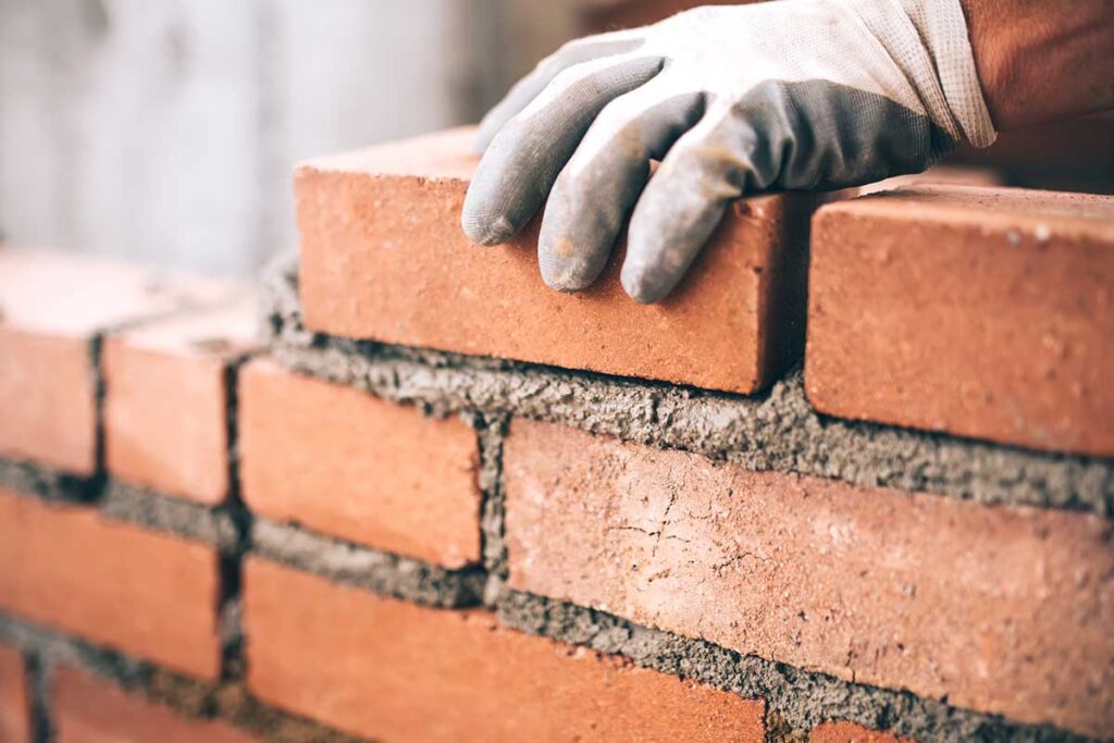 A bricklayer's gloved hand places a red-hued brick in place with mortar.