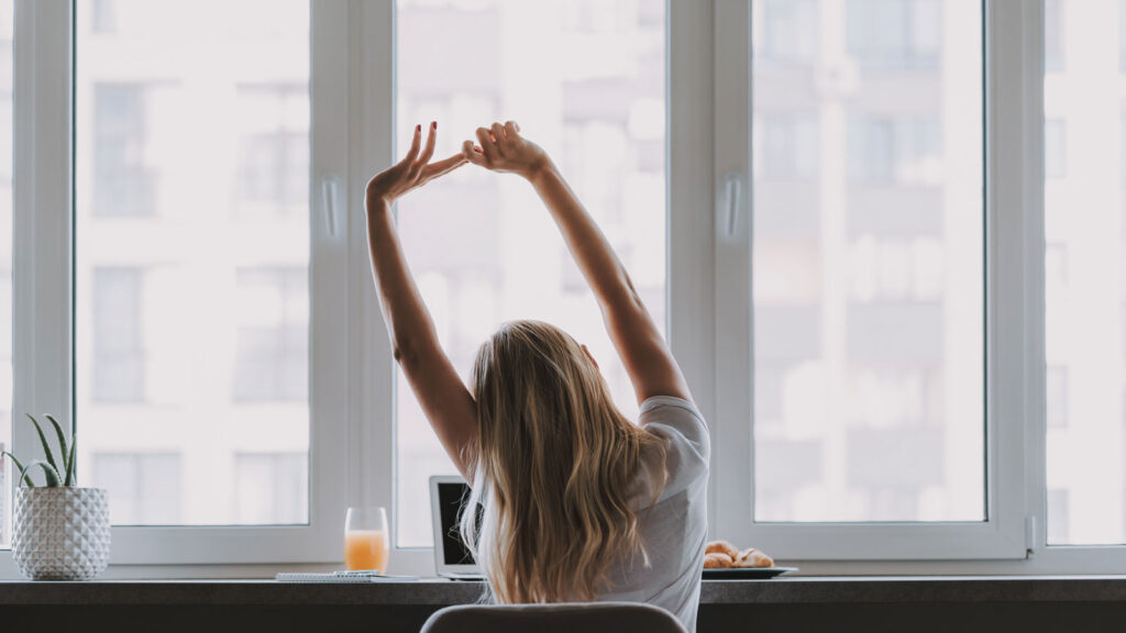 Back view of woman sitting at a desk stretching her arms over her head.