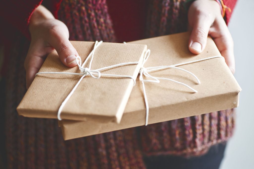 Two hands hold festive gifts wrapped in brown paper and tied with white string. The person is wearing a chunky, textured sweater in warm colors, perfect for the holiday season. The background is softly blurred.