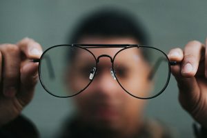 A photo illustration shows an out of focus individual in the background holding up rounded eye glasses for focus in the foreground.