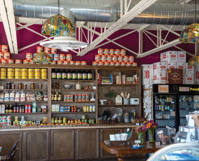 Step into a cozy store interior where wooden shelves brim with an array of canned goods and products. Colorful lamps hang above, casting a warm glow over the Stockyards experience. A refrigerated section beckons from one side, and a wooden table adorned with flowers completes the inviting scene.