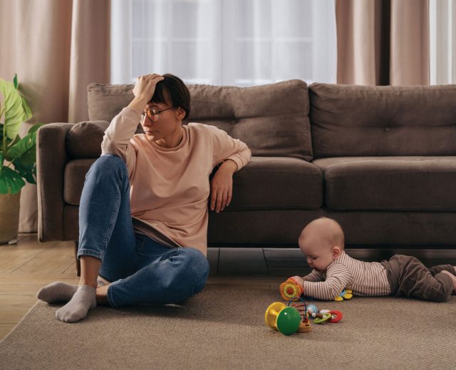 Woman-sitting-in-front-of-couch-on-floor-holding-her-head-while-a-baby-plays-nearby