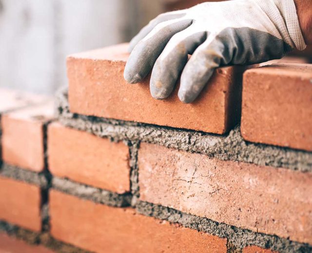 A bricklayer's gloved hand places a red-hued brick in place with mortar.