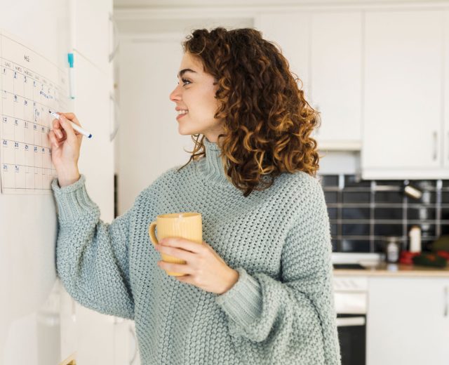 Woman with hot cup at home writing on wall calendar.